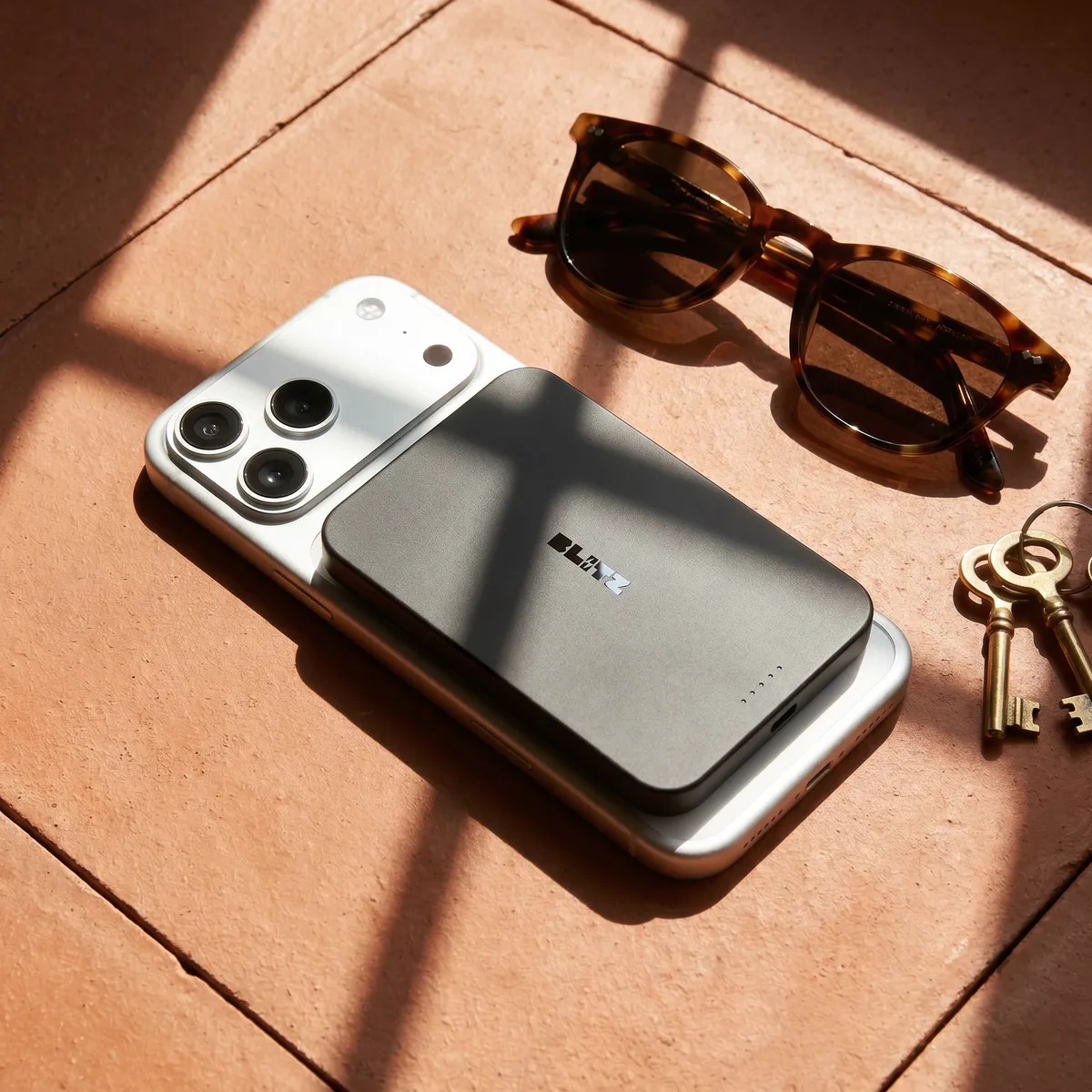 Black power bank on a phone in warm sunlight next to tortoiseshell sunglasses and keys