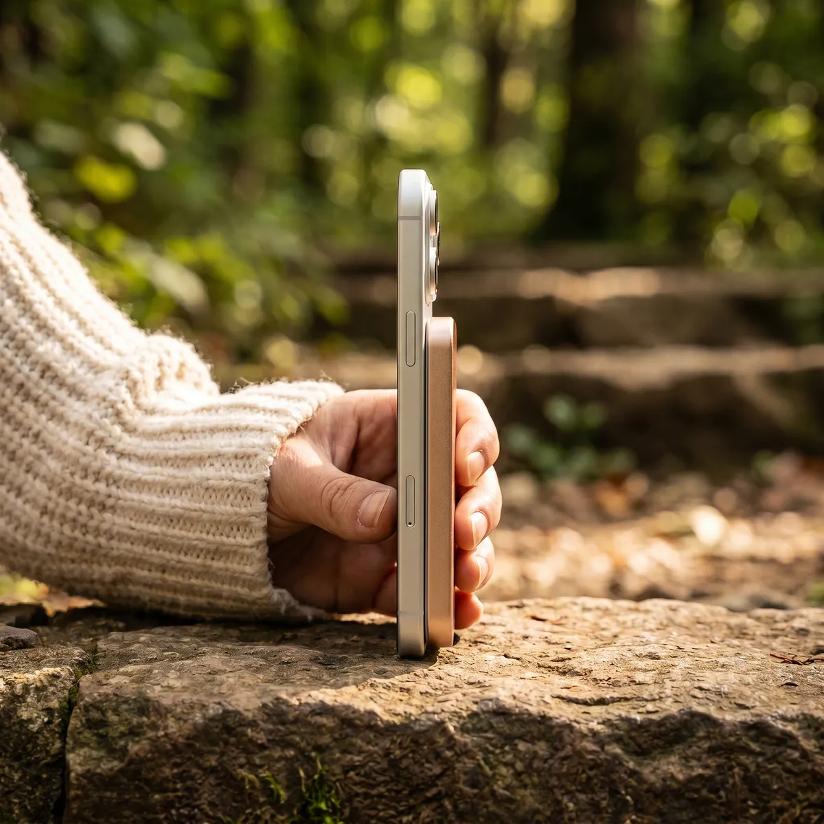 Phone with the rose gold power bank held upright on a mossy stone wall in a sunlit forest