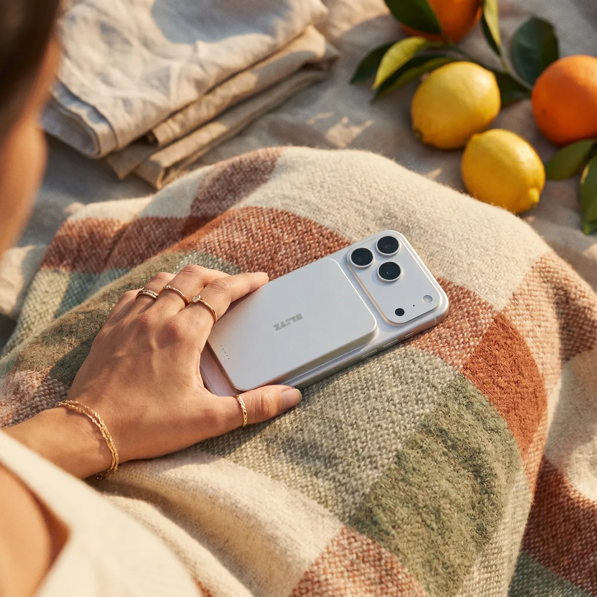 Hand with gold rings holding the silver power bank on a phone, on a checked blanket next to citrus fruit